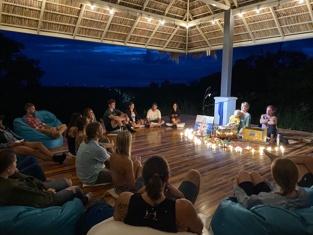 A kirtan circle gathering at dusk under a thatched palapa, candles arranged in the center.